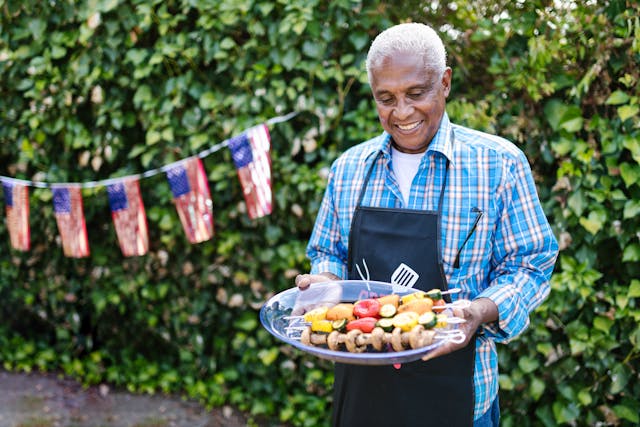 older man holding a plate of kebabs with USA flags in the background