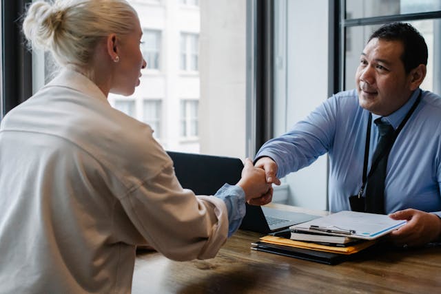 man in a dress shirt in front of a laptop and a checklist shaking hands with a woman across a desk